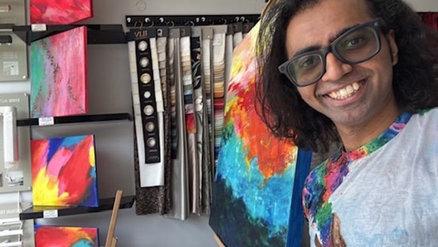 Vaibhav Sinha smiling at the camera, wearing glasses and a multicoloured T-shirt, in front of a large abstract painting in orange, blue and green. More brightly coloured canvases hang on the wall behind him.